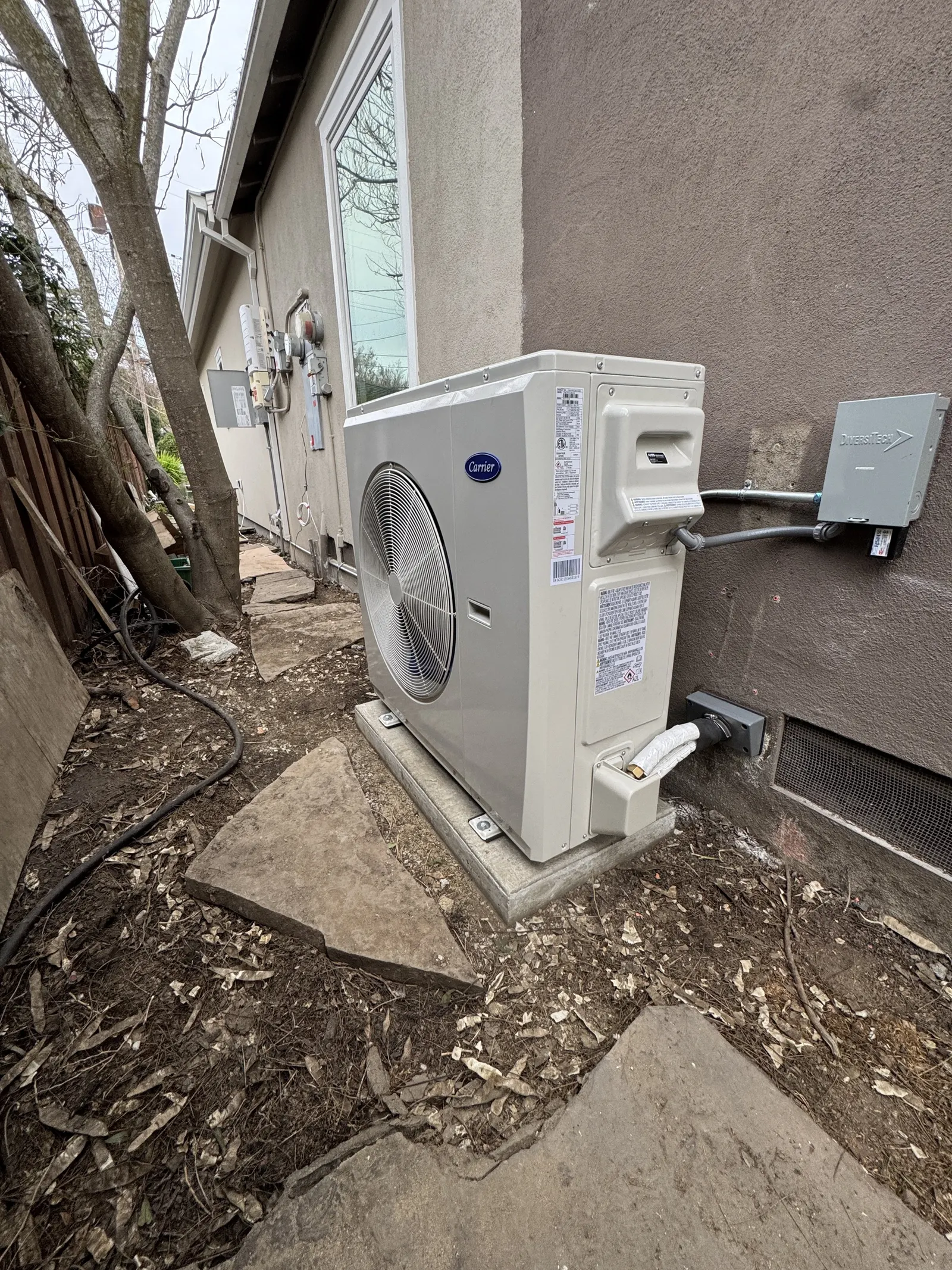Carrier outdoor heat pump on a low concrete pad next to a Pico-Robertson home with dedicated electrical disconnect and protected condensate line