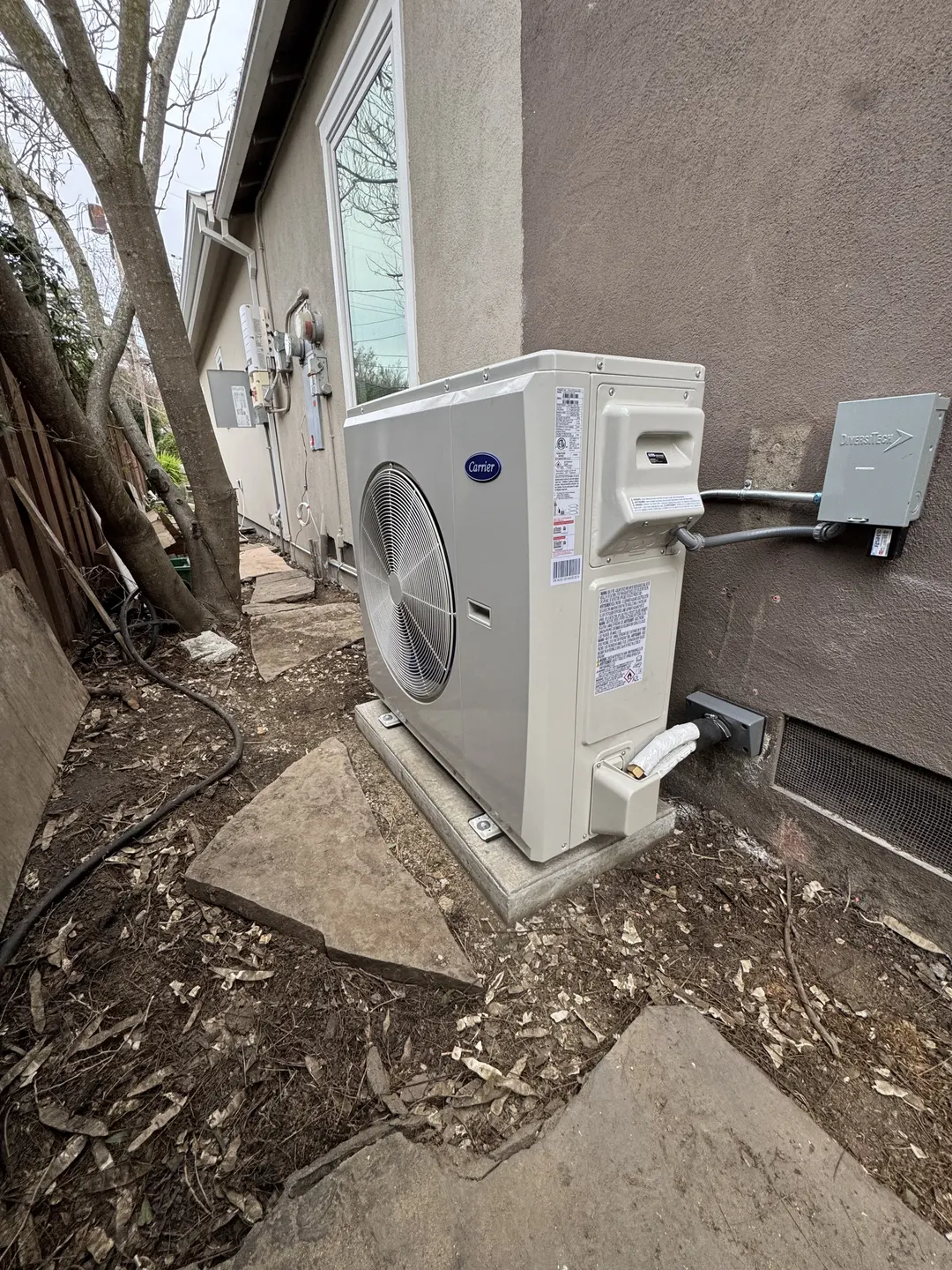 Carrier outdoor heat pump on a low concrete pad next to a Pico-Robertson home with dedicated electrical disconnect and protected condensate line
